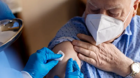 A nurse wearing blue clinical gloves administers a jab to the arm of an older man wearing a facemask