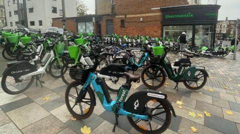 A street in London with dozens of hire bikes all over the pavement. A shop and a modern block of flats are in the background. 