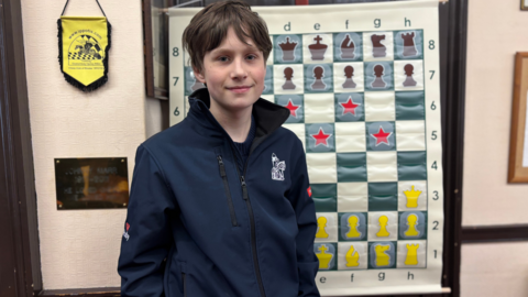 Frederick Waldhausen Gordon looking straight at the camera. He has dark hair. He is wearing a blue tracksuit top with a white logo on the upper right chest and is standing in front of a chessboard.
