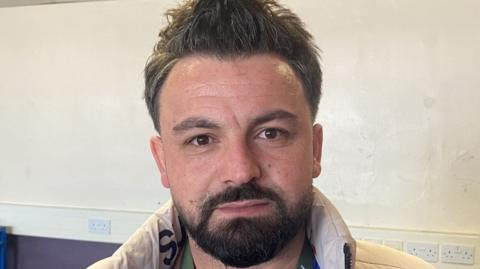 A man with short brown hair, beard and moustache with a cream jacket, looking directly at the camera