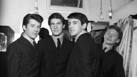 An old black and white image of the band The Searchers. They are standing together smiling wearing matching suits. 