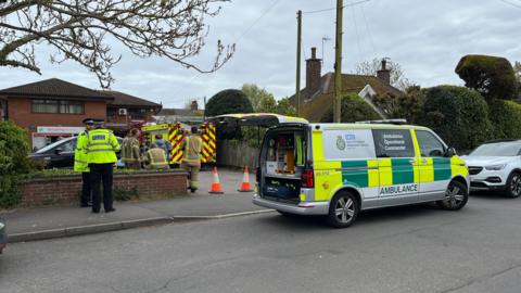 An Ambulance Operational Command vehicle is parked on the road outside the surgery with its boot open. Police officers stand nearby, there are orange cones blocking the entrance and a fire crew and engine in the car park.