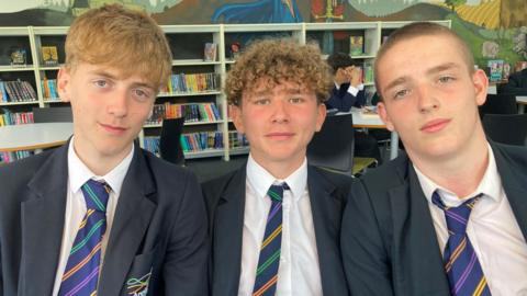 Three people seated close together in the foreground, facing directly toward the camera. All three individuals are wearing matching dark blazers, white shirts, and striped ties with diagonal bands in multiple colors, suggesting a school uniform. The person on the left has light-colored, slightly tousled hair, while the person in the middle has short, curly hair, and the person on the right has very short hair.
They are sitting in what appears to be a library or study area. Behind them, there are shelves filled with a variety of books arranged upright and in rows. The shelves are white and evenly spaced, creating a structured and organized background.