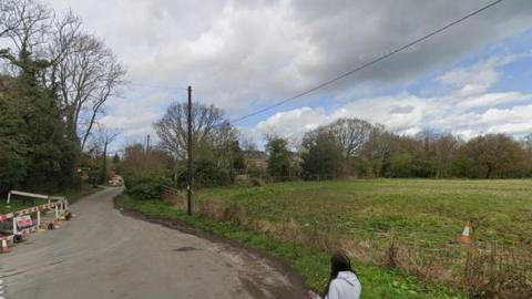 A field can be seen on the right hand side of a country lane, with trees and some houses in the distance.