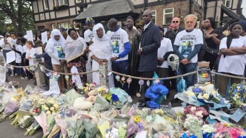 Ibrahima Seck's father clasps his arm around his wife while others stand beside them wearing white shirts with Ibrahima's picture. Floral tributes are laid out on the ground in front of them.