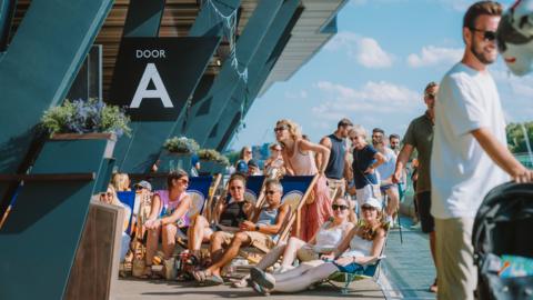 People sitting in deckchairs on an outdoor terrace under a sign reading 'Door A' at Fulham Pier on a sunny day.
