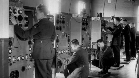 A group of men in military uniform standing in front of tall grey equipment bays. Some are kneeling to see the lower part of the display. Some are pressing buttons or rotating dials. There are shafts of light coming from windows behind the bays.