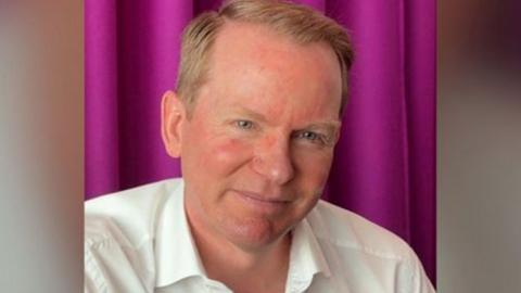 A man, Charlie Nunn, wears a white shirt. He has short red hair and sits in front of a purple curtain.