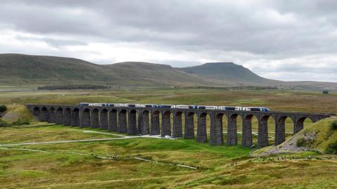 A train running along the Settle to Carlisle railway line over the Ribblehead Viaduct. The stone viaduct with its many arches is surrounded by rolling green land with fells in the background. 
