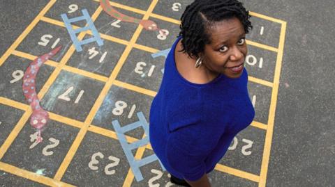 Carol, who is black, is stood on a snakes and ladders board, painted on the playground of Hunter's Bar Junior School. She has been photographed from above, so we are looking down on her - she looks up into the camera.