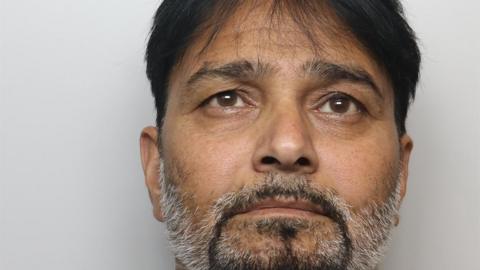 A mugshot of Qayum, who is facing directly toward the camera. He has short, dark hair. Facial hair is present, including a closely trimmed moustache and beard that extends along the jawline and chin, with grey hair visible within the beard.