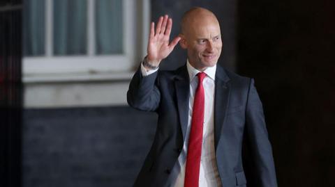 A bald man in a dark suit, white shirt and red tie.