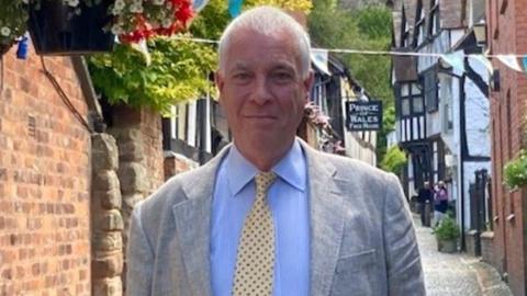 Ewen Sinclair is standing in a street in Ledbury, wearing a suit and tie - behind him are several black and white buildings.