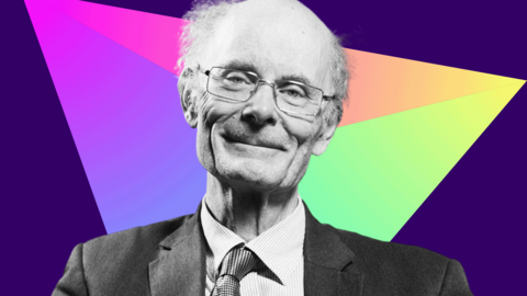 Black and white photo of Prof Sir John Curtice who has a bald head with grey hair at the sides. He is wearing metal-rimmed glasses, a dark suit and tie and a light-coloured shirt. He is smiling at the camera. The background is a prism r triangle made up of different bright colours
