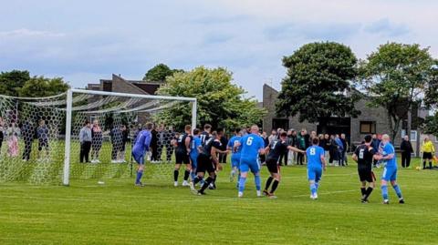 Kincorth in all black kits playing against Cowie, who are in blue. The players are in the box waiting for a corner to be crossed in. There's a row of spectators on the touchline, in front of trees. 