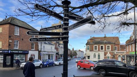 Beccles town centre with a black signpost in the centre, cars parked near shops and people walking around