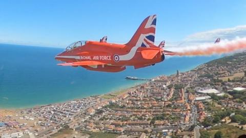 A small red jet with Union Flag livery on its tail flies over a coastal town, with the sea seen to the left of the image. The picture is taken from the cockpit of another plane flying alongside it.