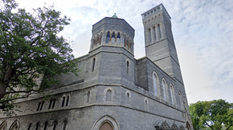 A Google maps image of the Plymouth Guildhall which is a large historic stone building. Trees seen in front on the left and behind on the right, with clouded skies behind.