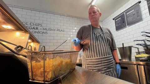 A man with short, ginger hair dips a basket containing chips into a fryer in a fish and chip shop.