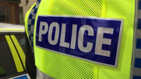 The back of a police officer wearing a bright yellow-high vis jacket stating 'police' in capitals in silver and blue. The side of a police car is on the left.