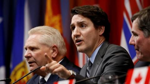 Canadian Prime Minister Justin Trudeau points and speaks into a microphone as he sits next to Ontario Premier Doug Ford. Provincial flags hang in the background.  