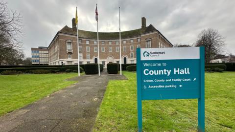 A turquoise sign reading 'welcome to county hall' in grass outside a curved brick municipal building with flags flying outside against a cloudy sky