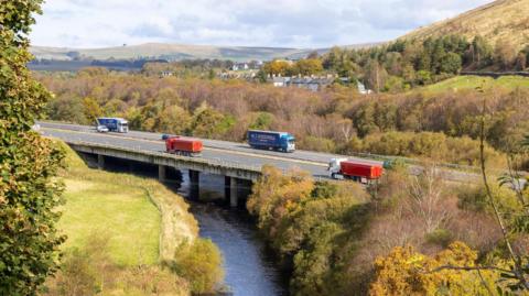 A high up view of the M6 motorway at the Lune Gorge between the Howgill Fells near Tebay, Cumbria with a number of lorries and cars travelling on the road in sunny conditions.