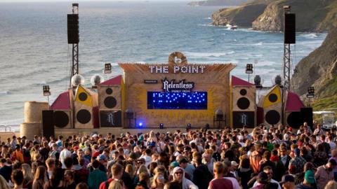 One of the sound stages called The Point at Boardmasters during the day with the crowd in the foreground and the sea and headlands behind it.