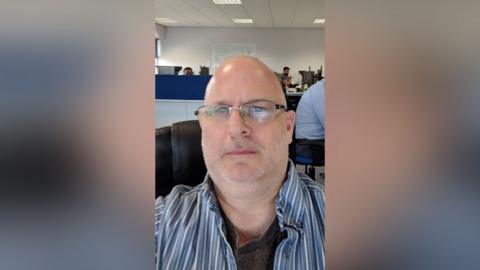 Steve Melrose has a shaved head, spectacles and is wearing a blue and white striped shirt with dark T-shirt underneath.