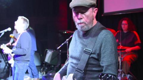 Three members of Blues Band DNA. Dave Penman is in the centre playing guitar he is wearing a cap and grey jumper and looking down. To his right is a band member in front of a microphone singing and to the right is a drummer. 