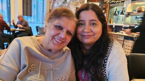 Rukhsana a woman on the right- with black hair, wearing a grey cardigan. Hugging another woman on the left, with light brown hair, wearing a cream colour shirt, smiling for the camera. They are sitting in a restaurant.