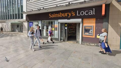 Google Street View image of people walking on the pavement outside a Sainsbury's Local shop on Sidwell Street in Exeter. Five people are walking past it. One of the people - a woman wearing a pink and navy blue rugby top looks like she is about to enter the shop. A cash point is on the exterior wall of the store, which has large windows on the front and double electric doors.