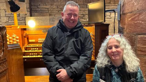 A grey-haired man, wearing a black jacket and a broad smile, is standing in front of the pipe organ. Alongside him is a woman with long wavy grey hair. She has a pair of red glasses on her head. 