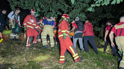 Various members of the emergency services some in red tops, others in blue tops with reflective stripes in a field in the dark, working together to rescue the walker, who is being carried on an orange stretcher.
