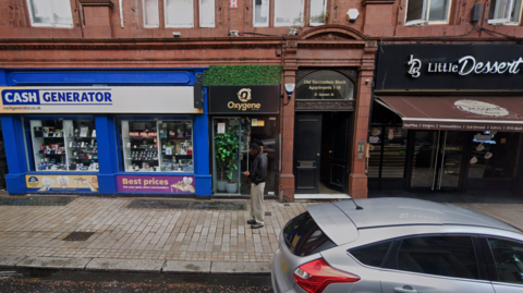 A man stands outside the doorway entrance to the club. There are shops either side of the premises. It has a black sign with yellow writing and artificial greenery arund it.