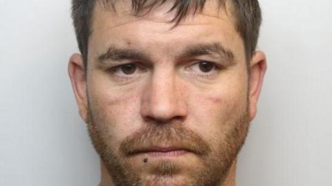 A custody image of Kane Thomas. He has brown hair and ginger beard and is looking off to the right of the image. 