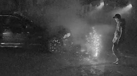 grainy footage of a man standing in a street while a car speeds around what appears to be a firework and a crowd gathered around them