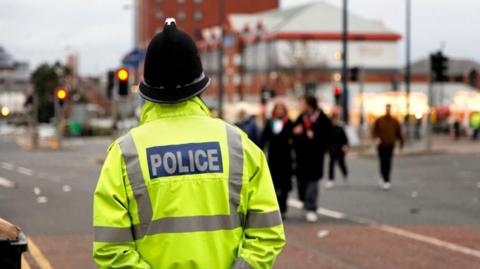 A police officer wearing a hi-vis jacket with "Police" on the back. They are stood in a street with people in the background.