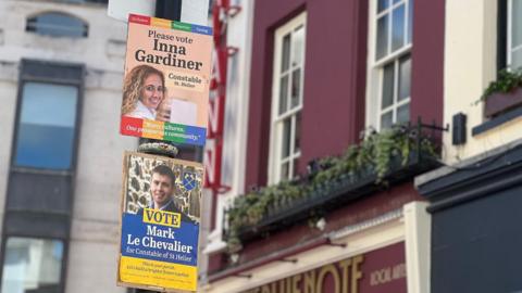 Two campaign posters are attached to a black lamppost on the street. The upper poster reads “Please vote Inna Gardiner, Constable St Helier” with a portrait and a slogan about community, while a second poster below promotes “Mark Le Chevalier for Constable of St Helier.” In the background are multi‑story buildings with red and cream facades, windows with flower boxes, and signs.