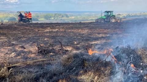 A picture of a smouldering moor, with some flames and smoke, with a tractor in the background on the right and some firefighters on the left.
