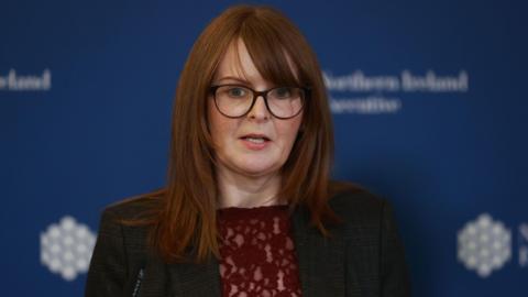 Caoimhe Archibald speaking at a microphone. She is wearing a grey blazer and red top and standing in front of a background with the logo of the Northern Ireland Executive