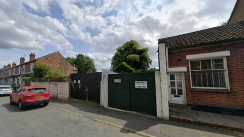 Two large gates mark out a space between two homes in the street. A red car is park outside one gate. No buildings can be seen behind the gates.