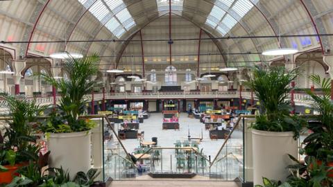 A view inside Derby Market Hall, looking down from the first floor to the ground floor. Plants are placed either side of a staircase with stalls visible in the background.