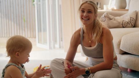 Becki Barratt is sitting on the floor in her living room next to her baby daughter Aréya, who is clapping her hands. 