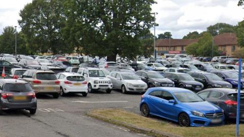 Lots of cars in a crowded car park with some trees and light brown buildings in the background