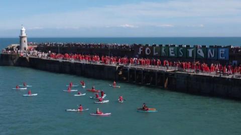 Many people in red dresses on a harbour arm. Canoe boats in the water.