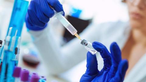 A woman wearing medical gloves in a lab holds a syringe in one hand and a small bottle in the other