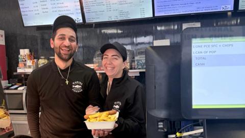 Both Natassa and Melios are wearing black baseball caps and black polo shirts with Marina Fish Bar written on them. Natassa is holding a takeaway tray with a sausage and chips in it towards the camera.