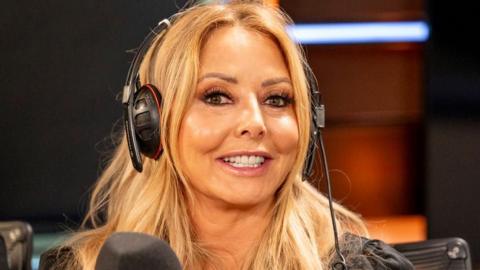 Carol Vorderman pictured in a BBC radio studio. She has long blonde hair and is smiling at the camera. She is wearing headphones, whilst sat in front of a microphone.