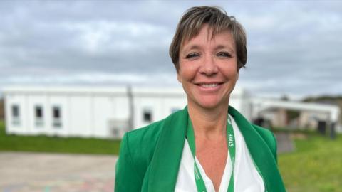 Jayne Crowley wears a green jacket with white blouse underneath, and a green staff lanyard around her neck. She has short cropped brown hair and is smiling at the camera. She stands in front of a white building with a patch of grass in front of it, next to a school playground.